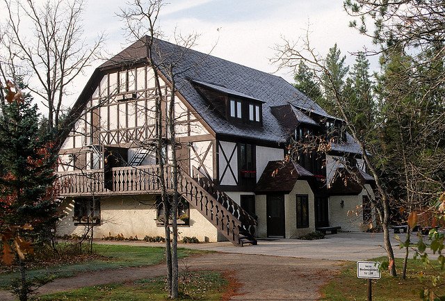 A german-style house with white paint and brown slats, standing among pine trees. 