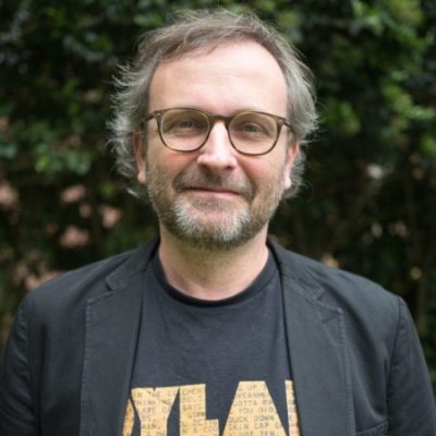 A grey-haired, bearded man wearing glasses and a black blazer over a t-shirt smiles at the camera.