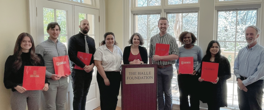 UGA dual degree students and ASHRAE fellow and director Dr. Thomas Lawrence stand in a slight arc around Dr. Heide Crawford, who is behind a dark wood podium that has a large gold plate etched with THE HALLE FOUNDATION in all capitals. The group has sunlit windows behind them, and every student is holding a red University of Georgia folder. 