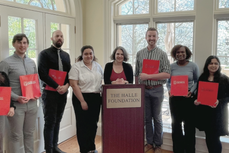 UGA dual degree students and ASHRAE fellow and director Dr. Thomas Lawrence stand in a slight arc around Dr. Heide Crawford, who is behind a dark wood podium that has a large gold plate etched with THE HALLE FOUNDATION in all capitals. The group has sunlit windows behind them, and every student is holding a red University of Georgia folder. 