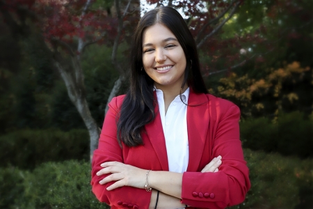 Mariah Cady - a caucasian dark haired young woman wearing a white button up and a red jacket. She is smiling. There are trees in the background. 